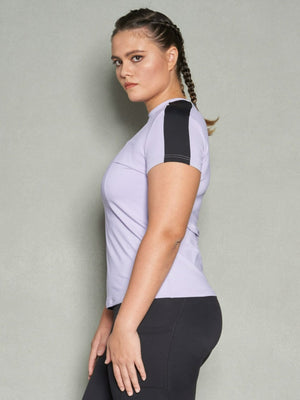 A half-body shot a woman with her body facing sideways while her face is looking at the camera. She is wearing a purple short sleeve women's workout top and black leggings.