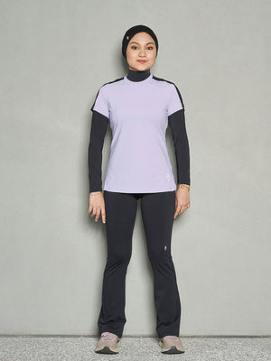 A shot of a woman standing straight looking at the camera while wearing a purple short sleeve women's workout top, long sleeve sports bra and black leggings.