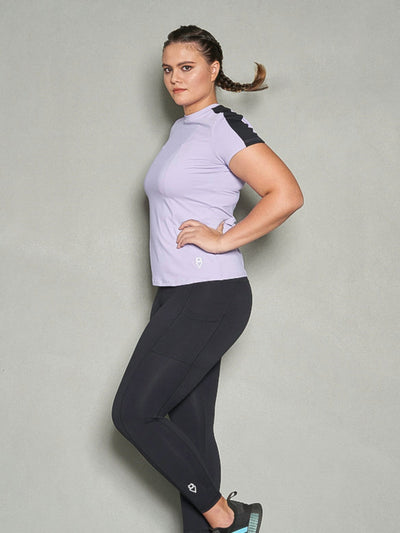 A shot a woman with her body facing sideways while her face is looking at the camera. She is wearing a purple short sleeve women's workout top and black leggings.