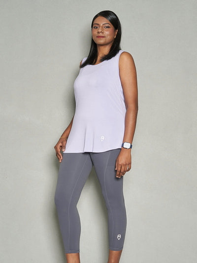 A shot of a woman with her body facing sideways while her face is facing the camera. She is wearing purple women's tank top and grey leggings.