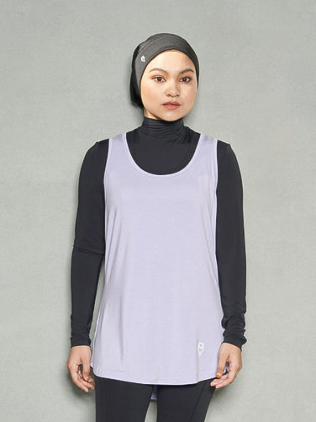 A woman standing straight facing the camera while wearing bandana sports hijab, purple workout tank top, long sleeve black sports bra and black leggings.