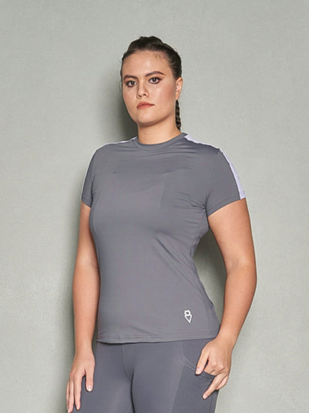 A half-body shot a woman with her body facing sideways while her face is looking at the camera. She is wearing a grey short sleeve women's workout top and grey leggings.