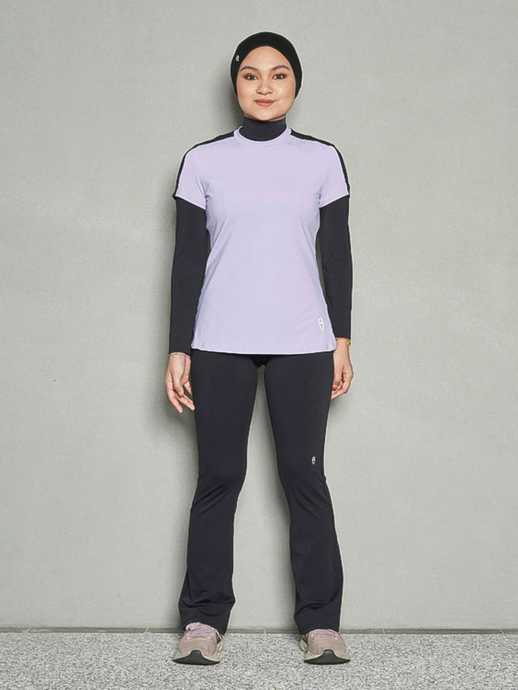 A shot of a woman standing straight smiling at the camera while wearing a purple short sleeve women's workout top, long sleeve sports bra and black leggings.