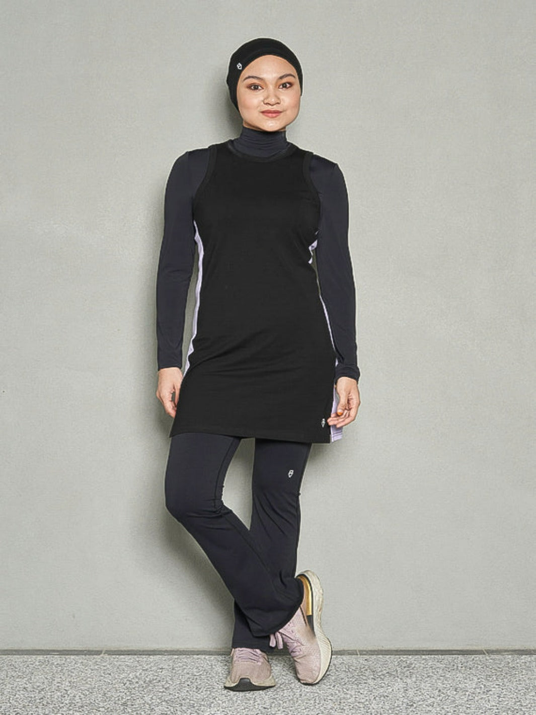 A shot of a hijabi woman with her face looking at the camera and her legs crossed. She is wearing bandana sports hijab, long sleeve sports bra, women workout top and bootcut pants.