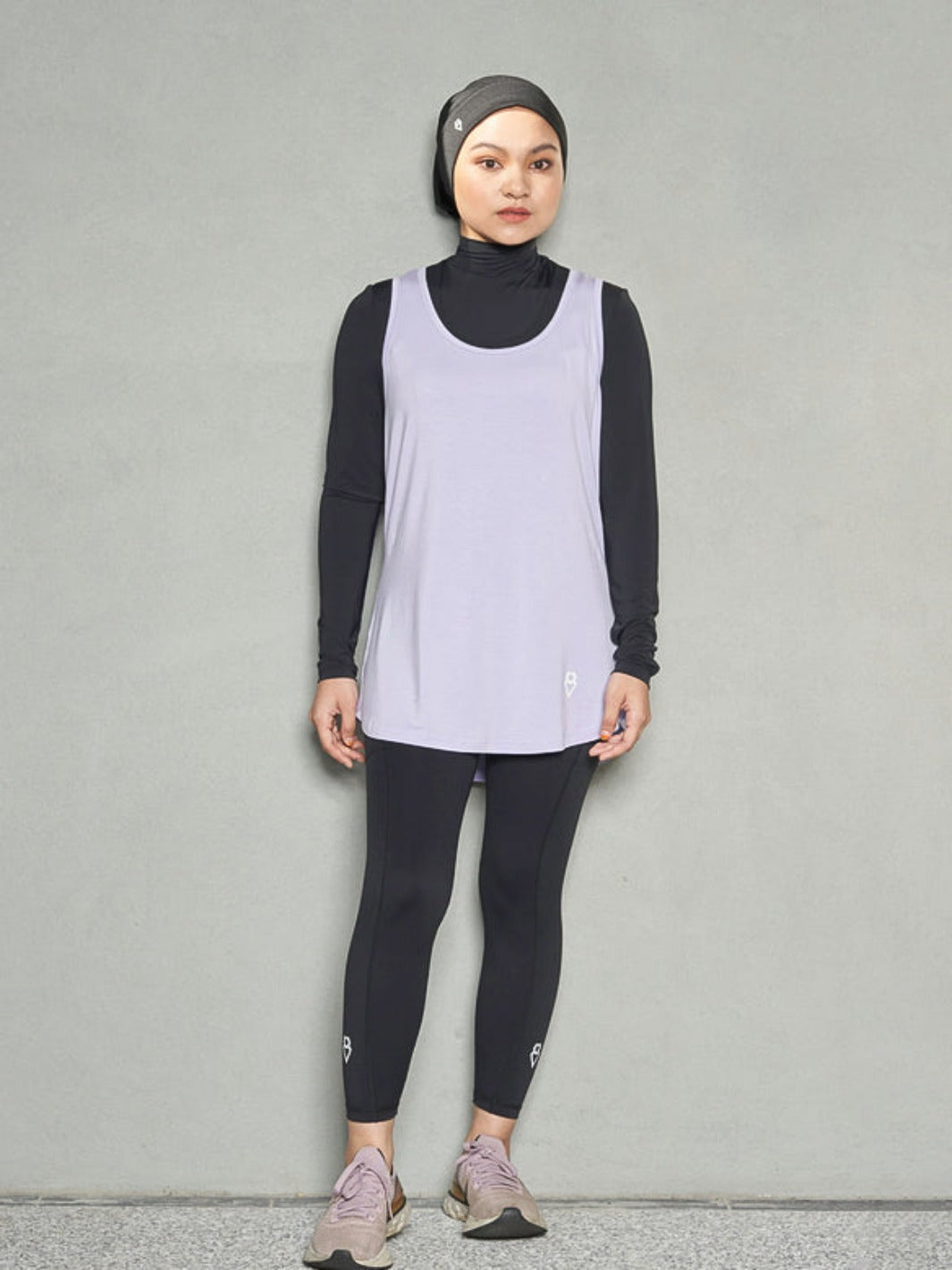 A shot of a hijabi woman standing straight looking at the camera while wearing bandana sports hijab, long sleeve sports bra, women's workout tank top and black leggings.