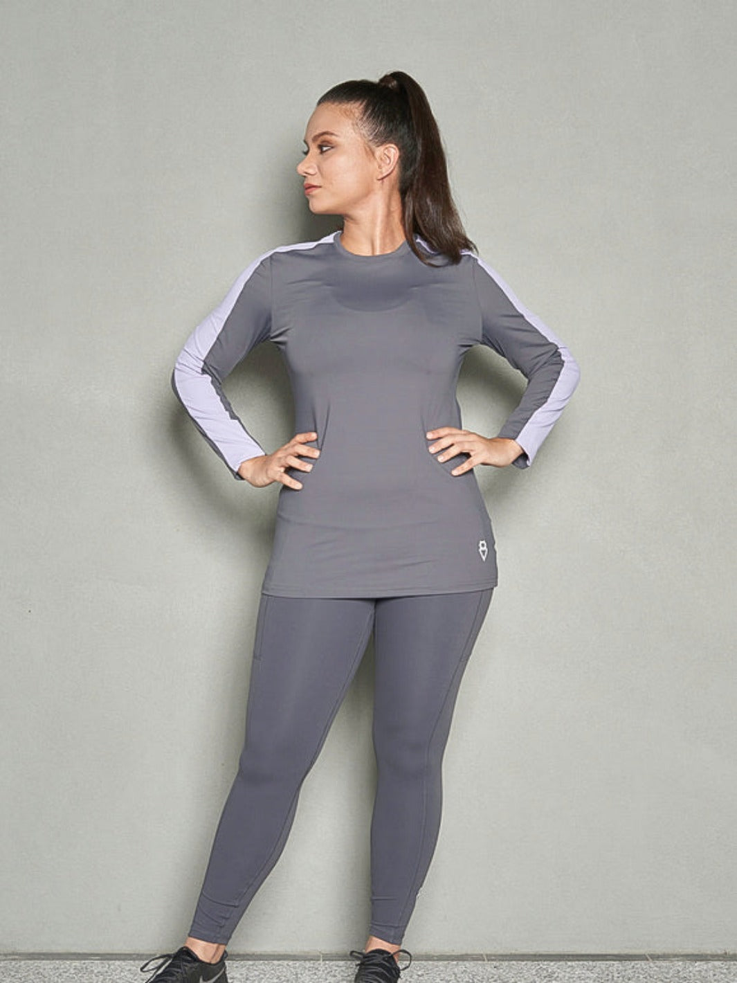 A shot of a woman looking sideways with her hands on her hips while her body is facing towards the camera wearing a grey long sleeve women workout top and grey leggings