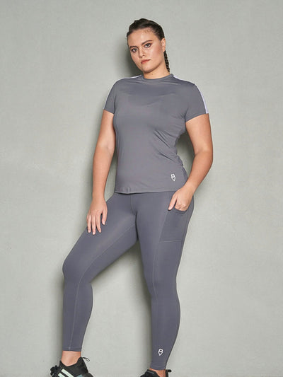 A shot a woman with her body facing sideways while her face is looking at the camera. She is wearing a grey short sleeve women's workout top and grey leggings.