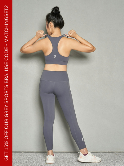 A shot of a woman from the back with both hands on the shoulder strap. She is wearing a grey sport bra and grey leggings.