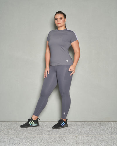 A shot of woman with her face facing towards camera and  her body facing sideways. She is wearing grey short sleeve women workout top and grey leggings.