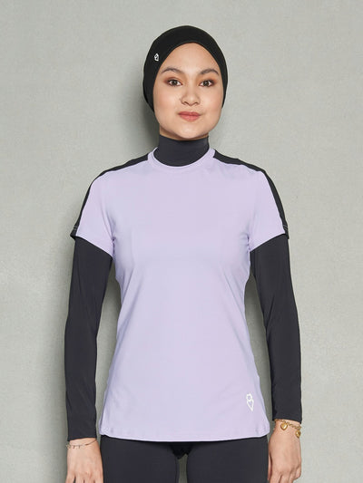 A half-body shot a woman looking at the camera while wearing a purple short sleeve women's workout top, long sleeve sports bra and black leggings.